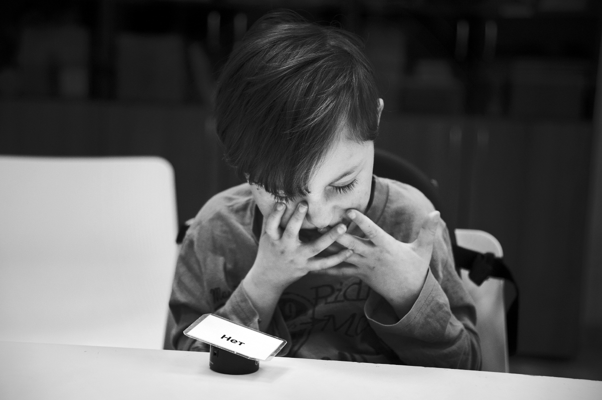 raisa mikhaylova . kids with special needs. Misha (7 y.o., cerebral palsy) sitting at the table with the special stuff helping him to answer the questions (It’s written "NO" there).